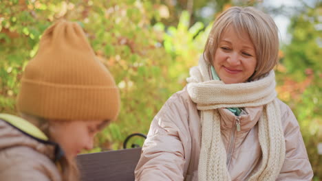 mother pulls fruit from coat pocket while child in orange beanie sits quietly beside her on wooden bench surrounded by golden leaves, warm smile on mother face reveals gentle care