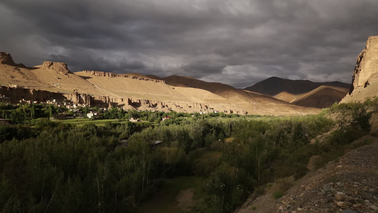 Canyon formation Mulbekh, swirling moody dark cloudscape over tree valley. Late afternoon dynamic golden light and shadows over royal cave monastery