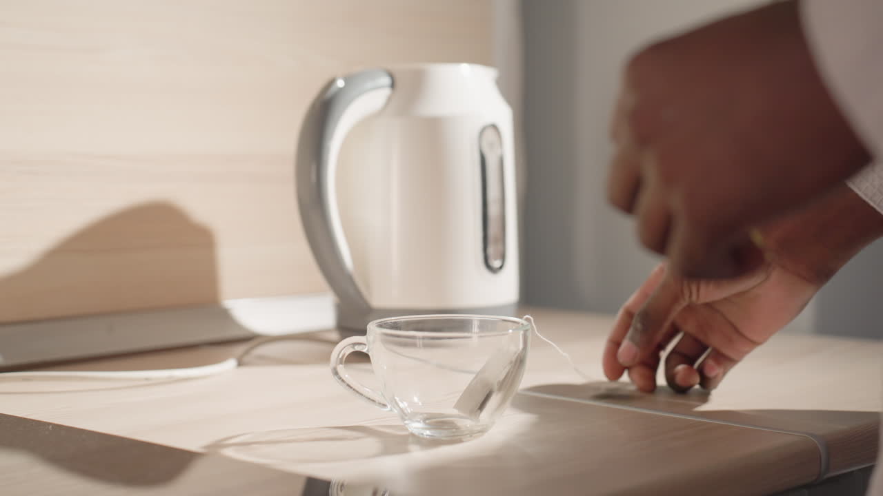 Close-up of man in bathrobe holding electric kettle above clear glass cup, preparing to pour water for tea or coffee. Tea bag in cup, soft morning light, modern minimalist kitchen in background