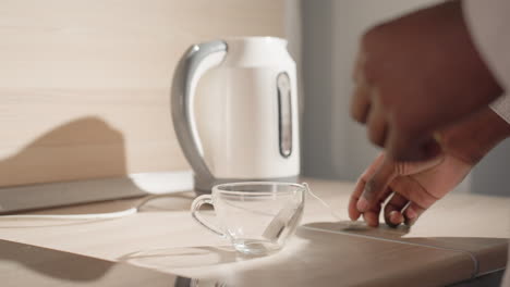 close-up of man in bathrobe holding electric kettle above clear glass cup, preparing to pour water for tea or coffee. tea bag in cup, soft morning light, modern minimalist kitchen in background