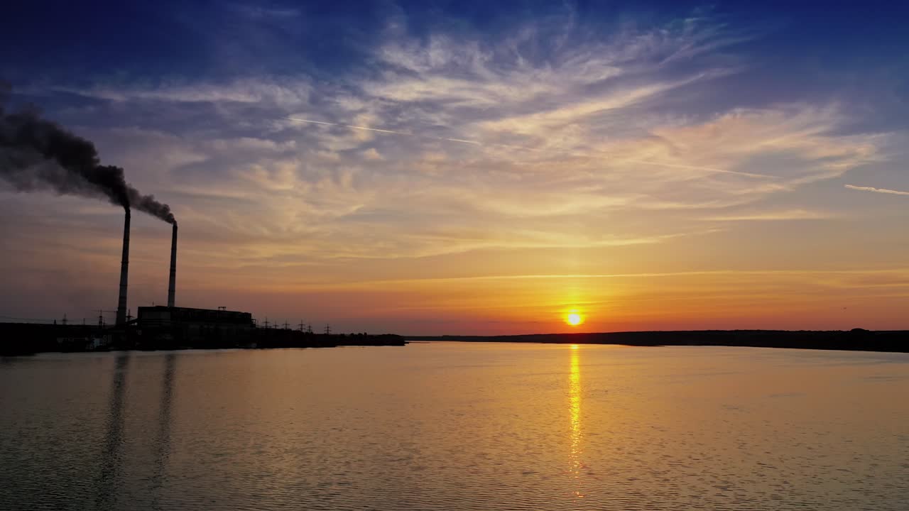 Orange sun reflects in water surface and industrial factory nearby. Beautiful evening background of setting sun on water. Manufacturing territory with smoke from pipes near the river.