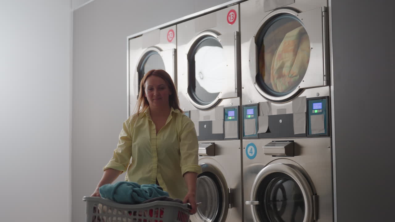 Work woman carrying laundry basket along laundromat row, industrial washers running with fabrics visible behind glass, stainless control panels, routine service, cleaning chores, indoor workflow
