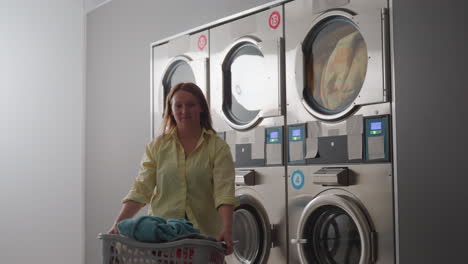 work woman carrying laundry basket along laundromat row, industrial washers running with fabrics visible behind glass, stainless control panels, routine service, cleaning chores, indoor workflow