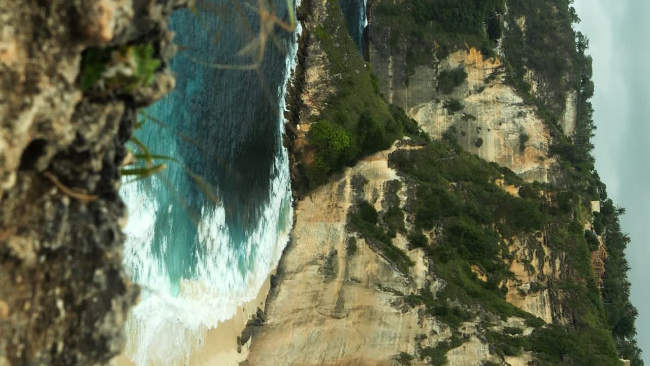 toma estática vertical de la playa de diamantes en la isla de nusa penida bali desde la cima del acantilado con olas rompiendo contra las rocas, y hierbas soplando en el viento en primer plano