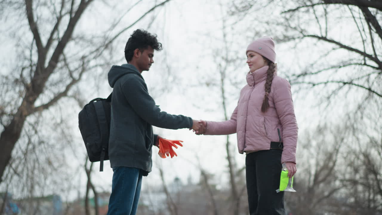 Black guy wearing dark gray hoodie holding bright orange gloves standing outdoors in park during early spring, bare tree branches in background under cloudy overcast sky, casual clothing and relaxed posture