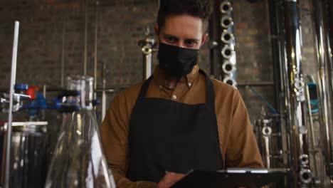 portrait of caucasian man working at gin distillery wearing face mask and apron looking to camera