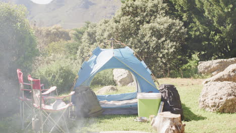 Smoking-firepit,-tent,-chairs-and-camping-equipment-in-sunny-countryside,-copy-space,-slow-motion