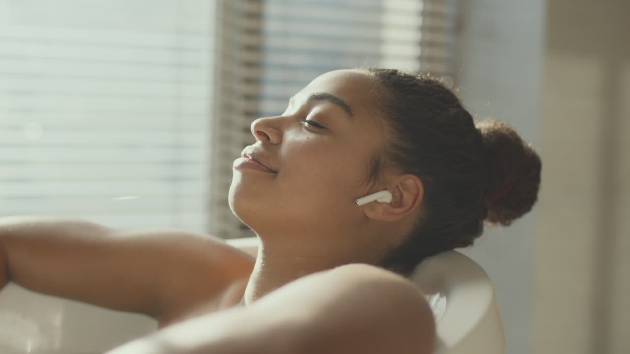 молодая афроамериканка отдыхает в ванне, слушает музыку с беспроводными наушниками, отслеживает выстрел