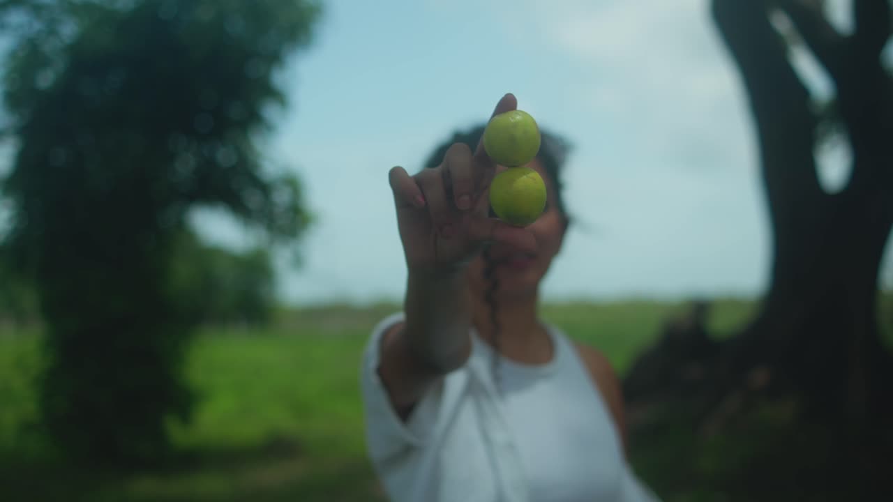 Soft-focus medium shot of young curly brunette woman framed by trees, gently moving two limes across frame, natural light