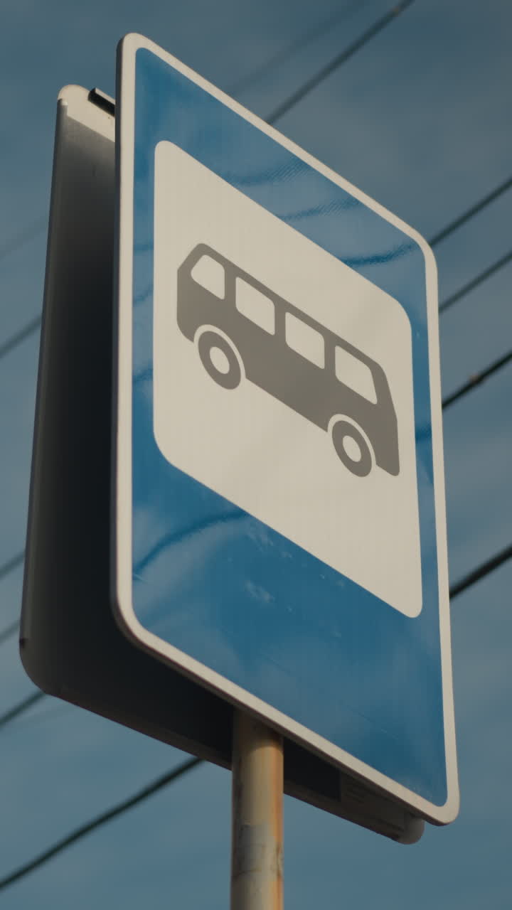 Señal de parada de autobús en poste oxidado, panel azul desgastado con icono blanco de autobús, cables aéreos contra cielo pálido, primer plano en ángulo que muestra superficie reflectante y pintura desconchada, detalle de infraestructura municipal