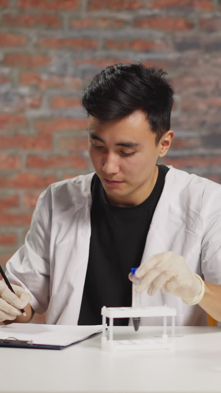 Young male Asian general practitioner in robe makes note on test tube with blood sample working at table with modern laptop in hospital laboratory