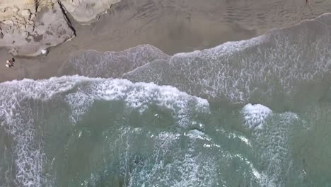 Aerial-view-of-rolling-waves-crash-on-a-beach-while-moving-upwards-to-reveal-more-sandy-beach-and-cliffs-with-people-laying-out-on-the-beach