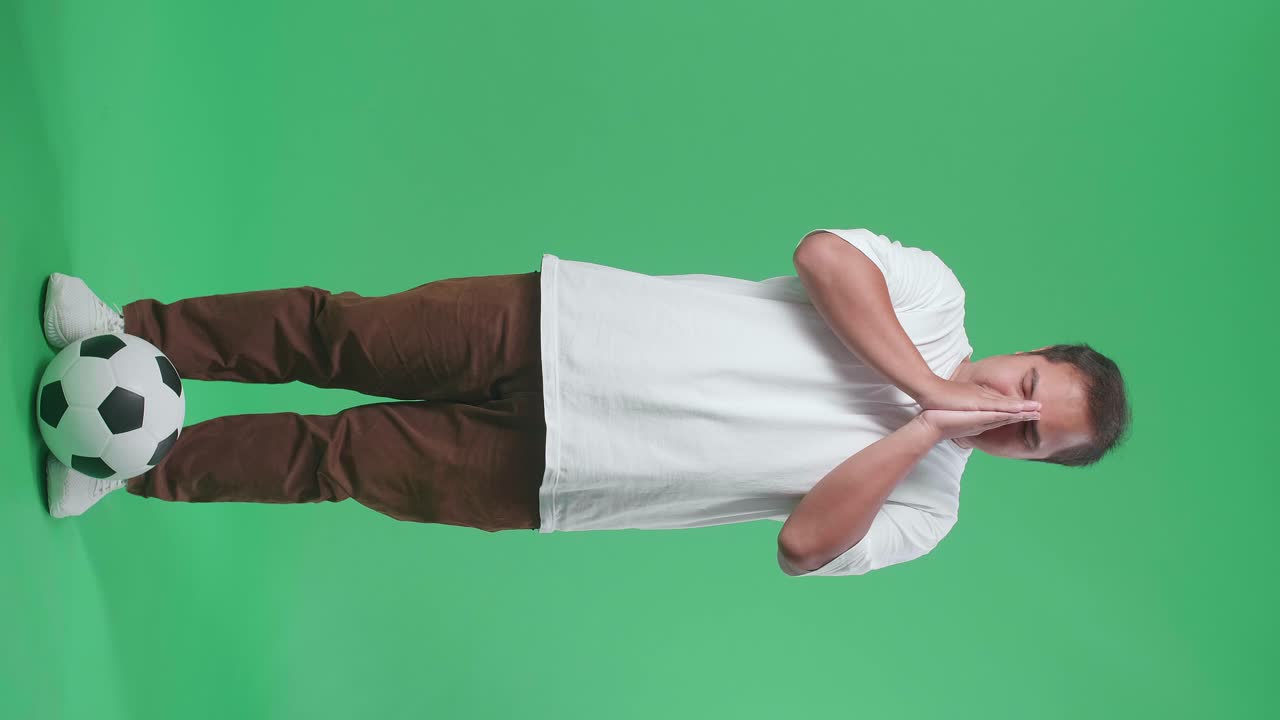 cuerpo lleno de ojos cerrados hombre asiático con una pelota rezar por un equipo antes de levantar la mano mientras aplaude el fútbol en el fondo de la pantalla verde