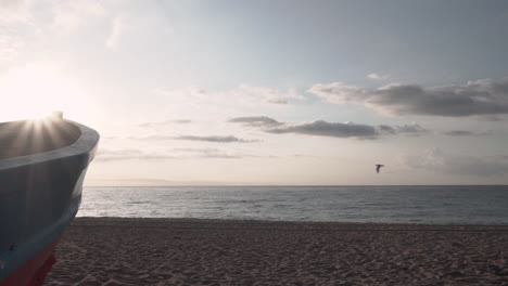 4K-Clip-Pfanne-Rechts-Von-Einem-Klassischen-Kleinen-Fischerboot-Auf-Dem-Sand-Eines-Strandes-Des-Mittelmeers-Bei-Sonnenaufgang,-Das-Einen-Sonnendurchbruch-Und-Einen-Vorbeifliegenden-Vogel-Zeigt