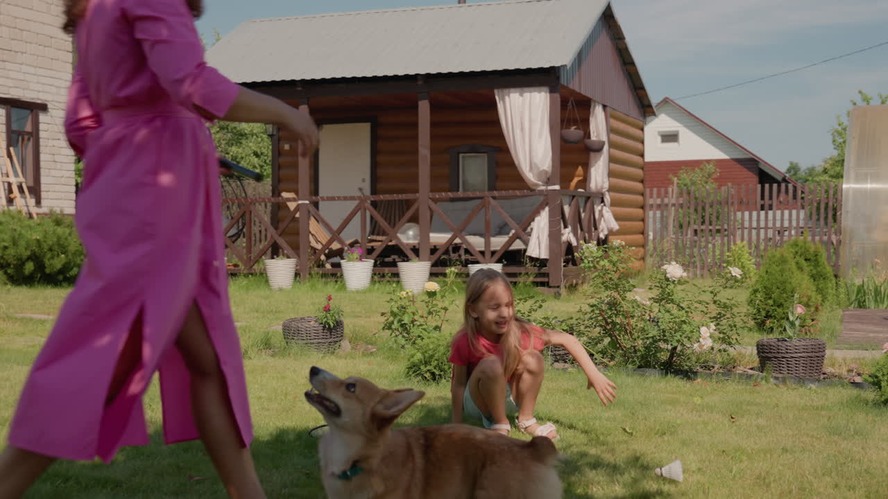 Sunny Backyard Caucasian Mother And Daughter Playing Fetch With Corgi Near Wooden Cottage, Cheerful Summer Afternoon, Lawn Games, Woman In Pink Robe Encouraging Child, Dog Chasing Ball, Relaxed Family