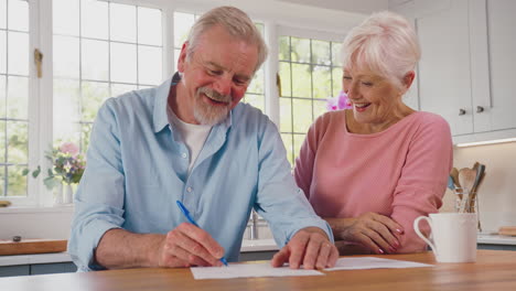 Smiling-Retired-Senior-Couple-Sitting-In-Kitchen-At-Home-Signing-Financial-Document