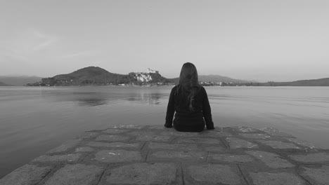 Back-view-of-child-girl-sitting-on-jetty-edge-facing-Angera-castle-on-Maggiore-lake,-Italy