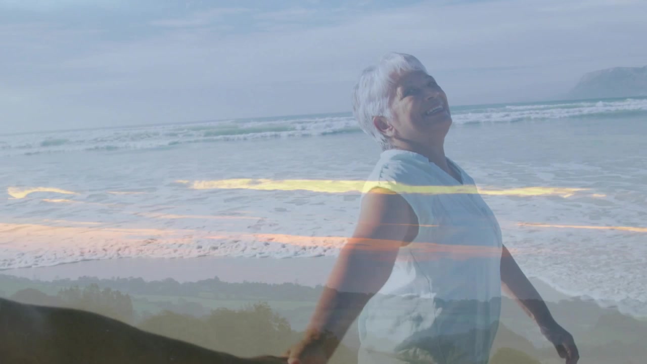 Coppia anziana che cammina sulla spiaggia, il partner le tiene la mano per darle stabilità, la donna si inclina sorridendo sotto il tramonto