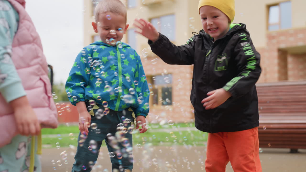 Excited boy and girl standing near bubble machine surrounded by floating soap bubbles, smiling happily during playful outdoor moment on playground, enjoying carefree childhood fun