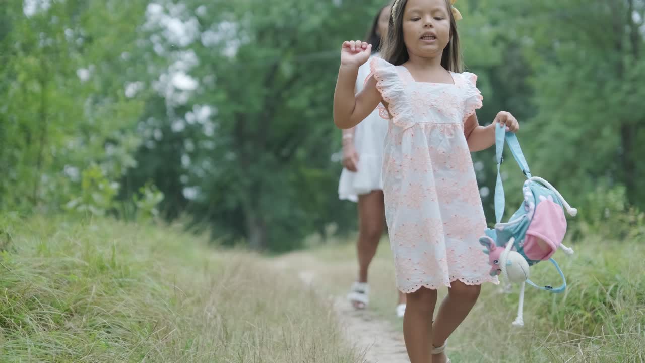 una mujer embarazada y su pequeña hija, ambas vestidas de blanco, caminan de la mano a través de un parque cubierto de hierba. la escena captura un momento sereno de unión familiar en la naturaleza, rodeada de árboles.