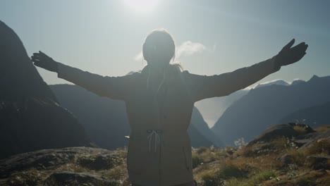 female hiker in alpine landscape with bright sunshine behind raising arms in happiness