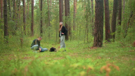 tourists in forest as man leans down to check green bag while woman stands nearby observing surrounding woodland quietly, bags on ground, surrounded by tall trees and lush greenery