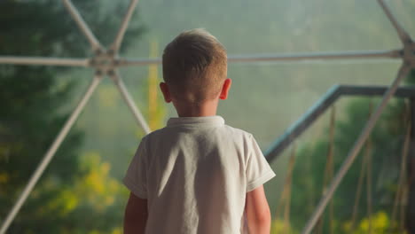 kid approaches window looking at landscape. child crosses arms on chest fantasizing slow motion on blurred background backside view closeup