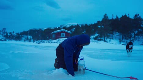 un hombre, junto con su perro, recoge agua fría de un agujero en el lago congelado en bessaker, condado de trondelag, noruega - toma estática