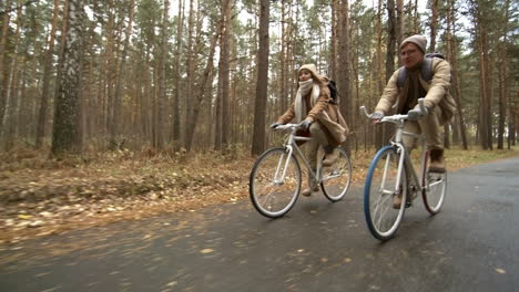 Side-View-Of-A-Couple-Wearing-Winter-Clothes-Riding-Bikes-In-The-Forest-While-Raining