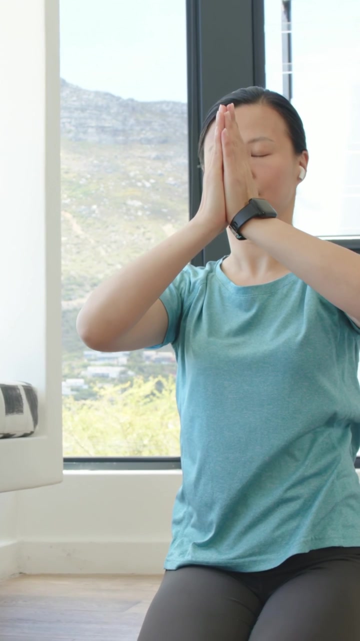 縦型動画：息を吸い込みながら、ティール色のTシャツとレギンスを着たアジア人女性が、落ち着くために窓の近くで腕を上げている。
