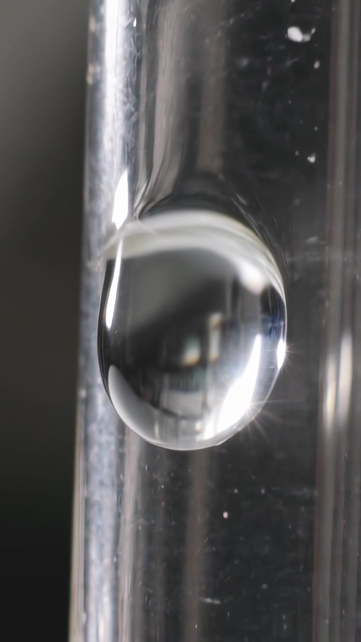 Video verticale: Gocciolina aderente che cede sotto la gravità in un tubo di vetro, con riflessi e polvere che si spostano