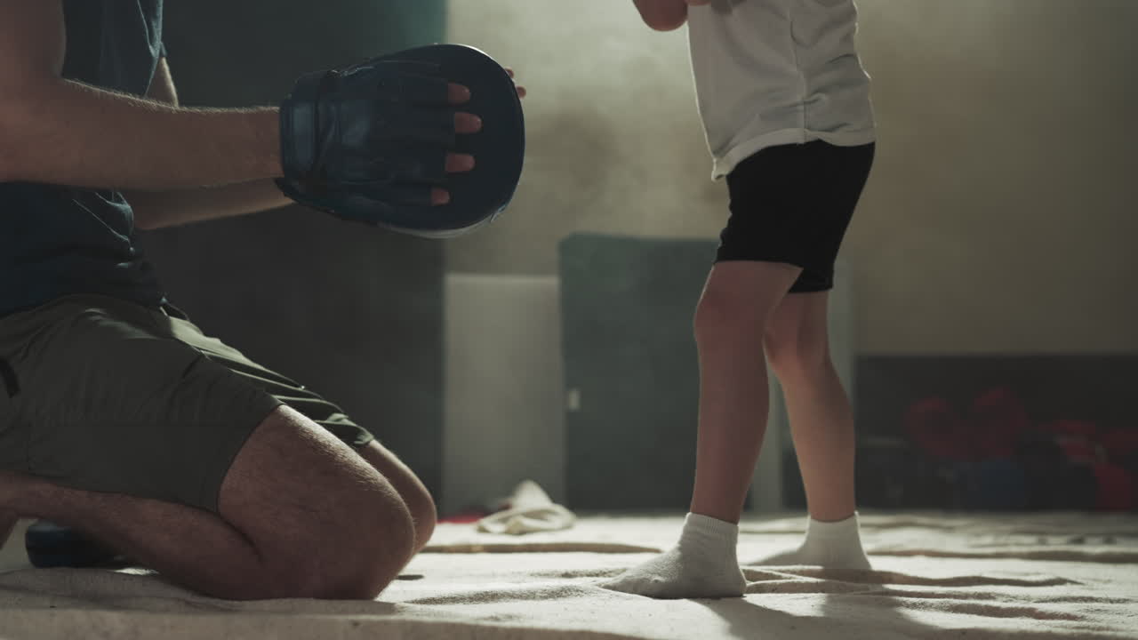 Kid kicks pad on trainer hand in martial arts gym. Skilled trainer and toddler boy study hits by legs in combat center slow motion. Kickboxing fight