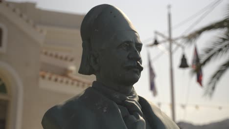 Close-up-of-saint-nicolas-statue-in-front-of-Agios-Mamas-Church-in-Spetses-greece-with-palms,-flags-and-the-church-in-the-background-as-a-symbal-of-the-ottoman-war-and-armata