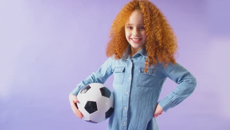 studio shot of young girl holding soccer ball under arm against purple background