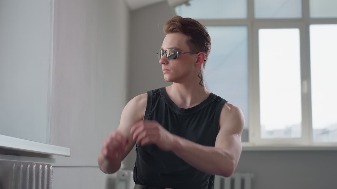 Man with neck tattoo seated by tilted window examines dark shades before putting them on, soft daylight illuminating profile and angular architecture while he strikes stylish contemplative pose