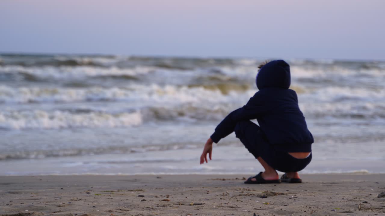 Boy resting near the sea at sunset. Child is painting on sandy beach on foamy waves background of the evening sea.