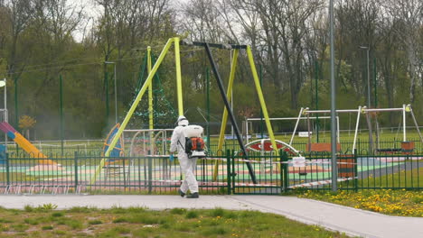 disinfecting public space and children outdoors playground. man in protective suit spraying with spray gun
