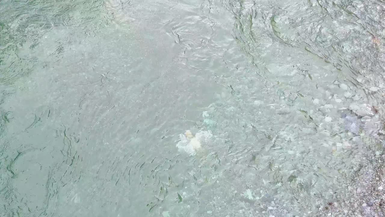 Aerial view of transparent, fast-moving stream water over smooth stones and pebbles in natural daylight. Subtle camera movement follows the river’s gentle current