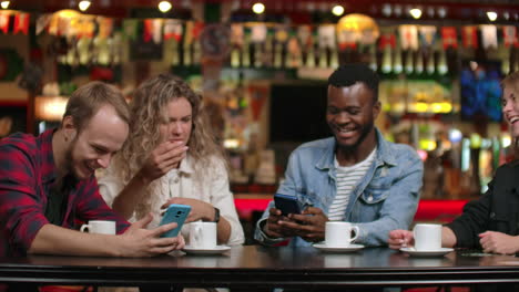 Vier-Freunde-Sitzen-In-Einem-Café-Und-Lachen-Und-Lächeln,-Während-Sie-Auf-Die-Bildschirme-Ihrer-Telefone-Schauen-Und-Reisefotos-Austauschen.-Afroamerikanische-Freundin-Plaudert-Und-Trinkt-Kaffee.-Multinationales-Unternehmen-Von-Freunden.