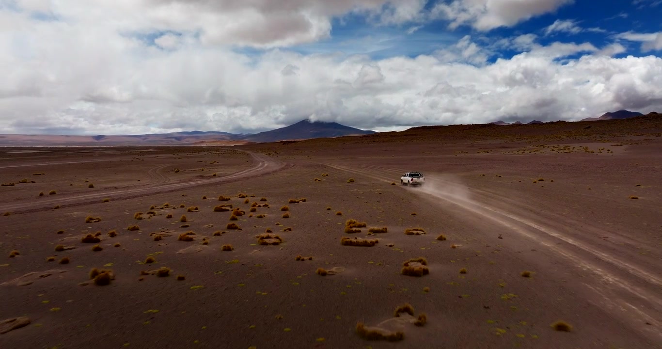 Veicolo fuoristrada che attraversa il deserto di Siloli in Bolivia sotto nuvole drammatiche, ripresa dinamica posteriore con drone che segue attraverso pianure vuote mentre la polvere si alza.