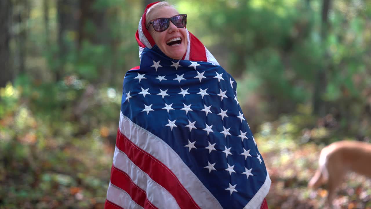 mujer rubia levantando una bandera americana detrás de ella con expresión sonriente y envolviendo la bandera alrededor de su cabeza y cuerpo