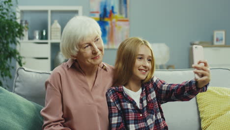 Close-Up-Of-The-Cute-Teen-Girl-And-Her-Smiled-Gray-Haired-Grandmother-Posing-To-The-Smartphone-Camera-While-Taking-Selfie-Photo-On-The-Couch-At-Home