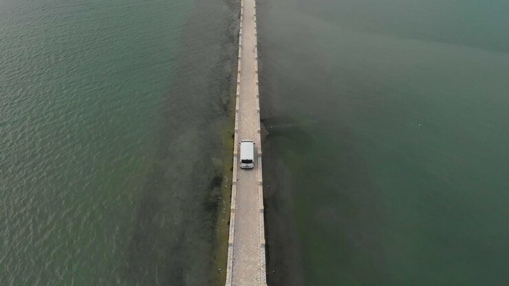 Riprese aeree con drone che seguono un furgone bianco mentre attraversa una strada su un ponte, arrivando a una piccola isola esotica greca, Koukoumitsa Vonitsa, Grecia.