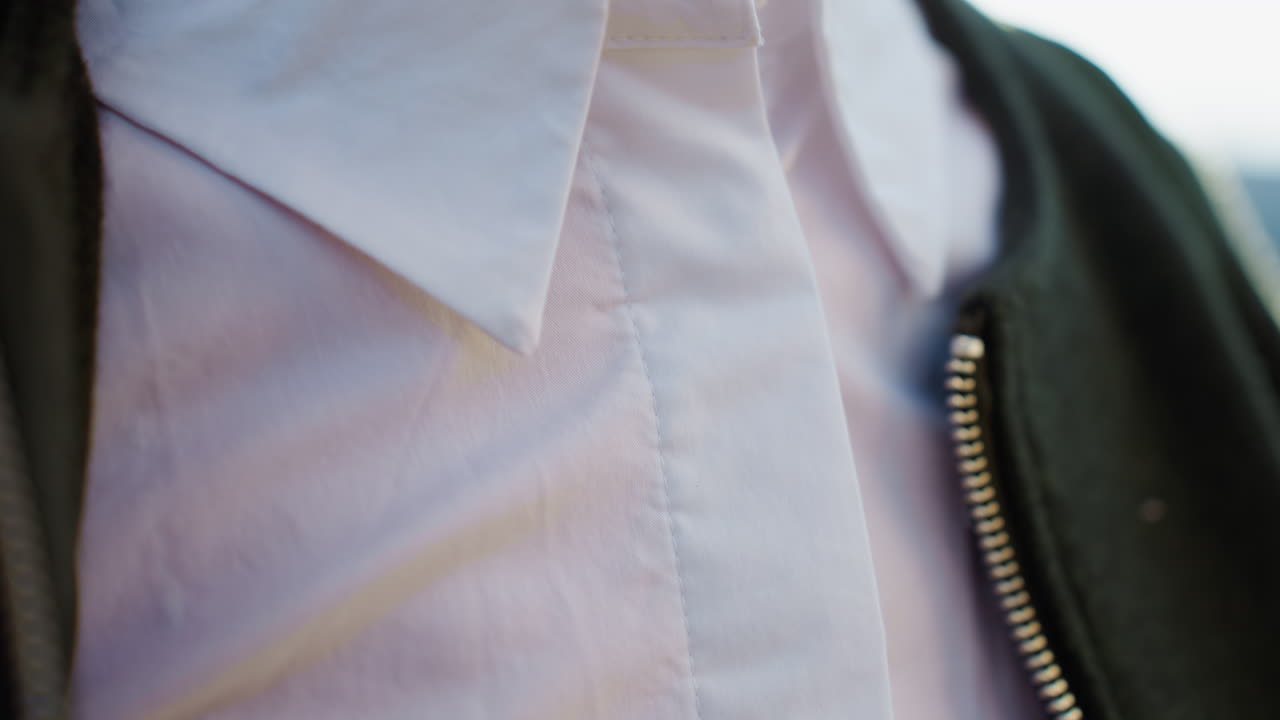 Close up adult woman zipping dark jacket outdoors, red polished nails visible, hands adjusting zipper near collar over white shirt, natural sunlight highlighting detail of casual clothing and gesture