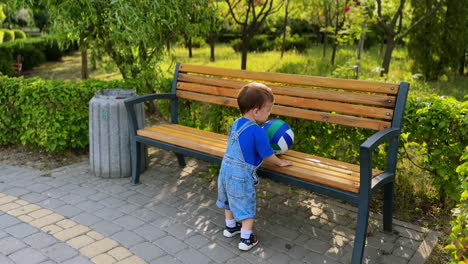 following the cute baby boy carrying a ball in the park. little kid comes up to the bench, puts the ball on it, watches it fall and picks from the ground.