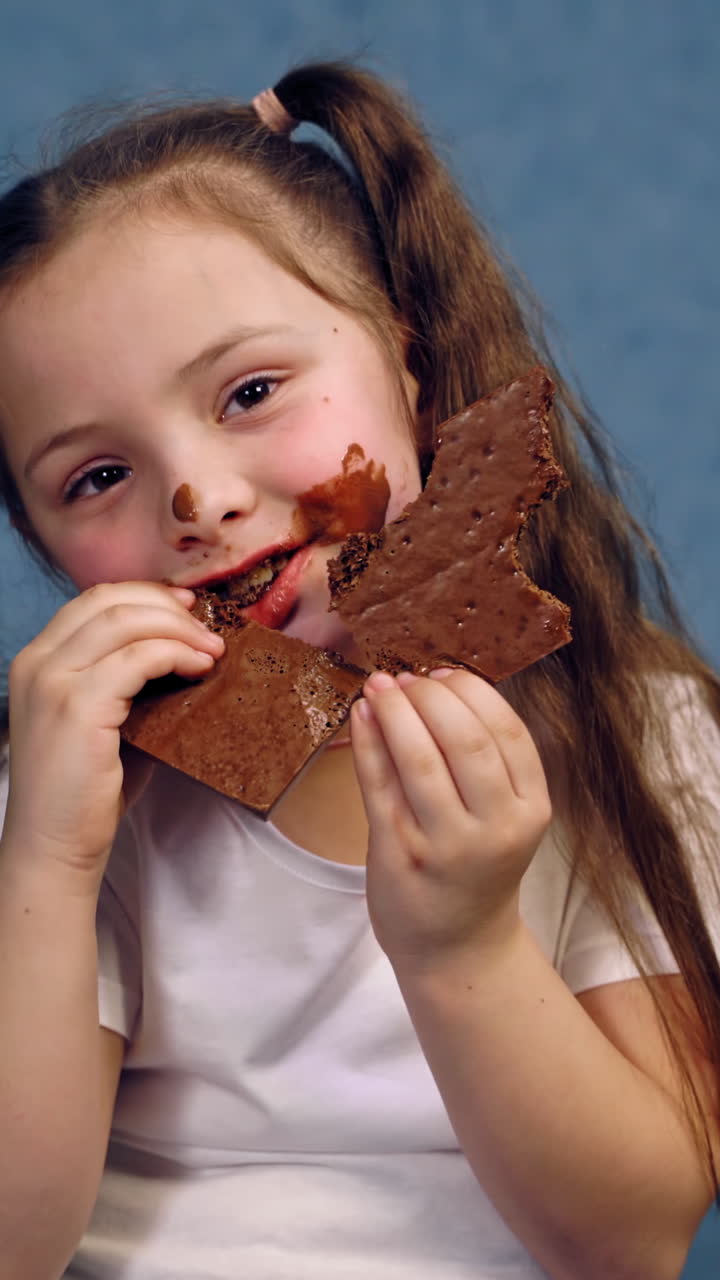 Portrait of a little girl eating chocolate. Funny kid with ponytails eats sweets with smeared face. Dirty child on blue studio background. Vertical video