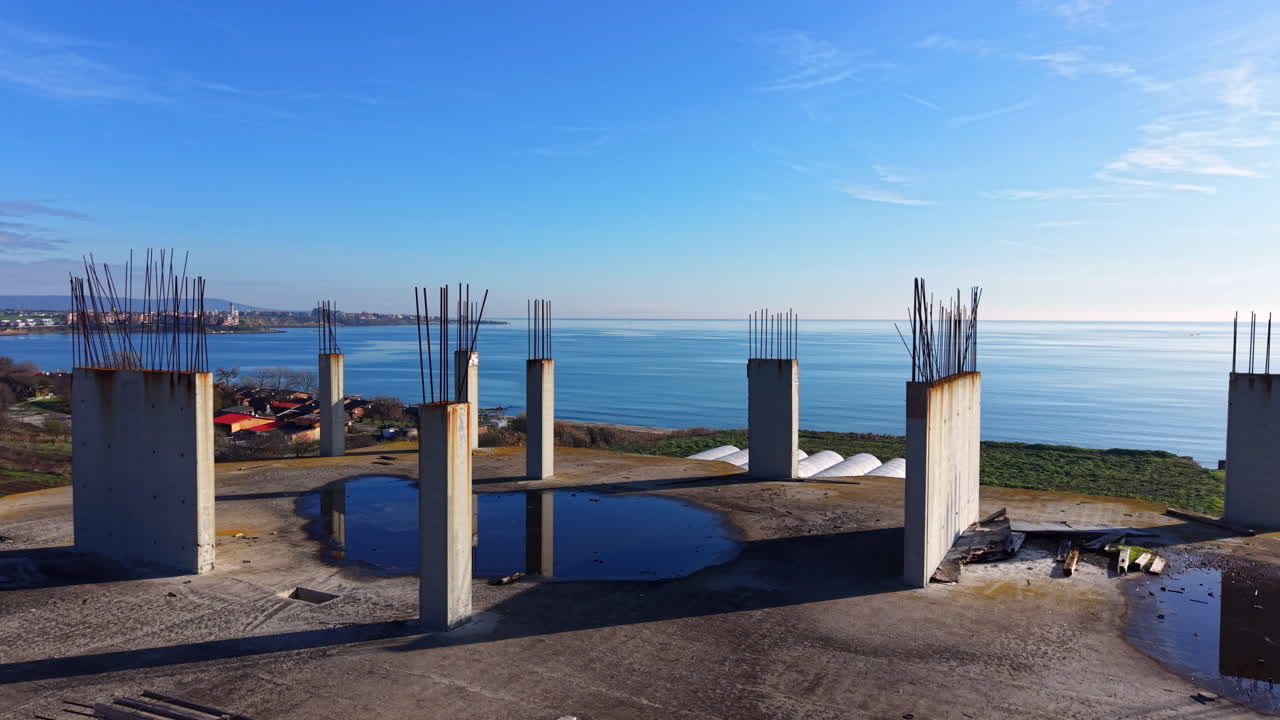 Ampia veduta di un grande tetto in cemento con numerose colonne verticali. Le pozzanghere riflettono il cielo blu. Mare e costa visibili sullo sfondo. Vista aerea