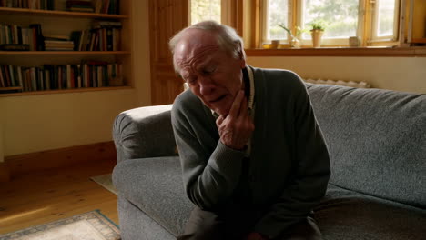 elderly man experiencing pain or discomfort in his throat or jaw while sitting on a sofa at home