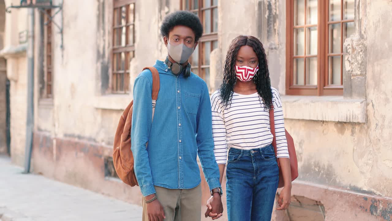 Coppia afroamericana con mascherine che si tengono per mano mentre guardano in camera per strada.