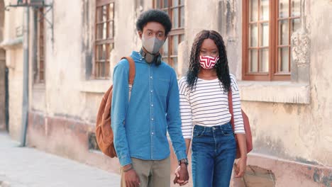 pareja afroamericana usando máscaras y tomándose de la mano mientras miraba la cámara en la calle